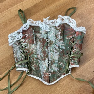 Blommig korsett med spetsdetaljer - Säljer en superfin korsett med blommigt mönster i gröna och bruna nyanser. Den har gröna snören på sidorna och baksidan för justering, samt vackra vita spetsdetaljer längs överkanten. Perfekt för fest eller speciella tillfällen!