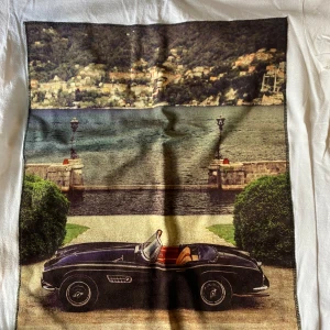 Vit t-shirt med biltryck från Bastille - Säljer en vit t-shirt från Bastille med ett coolt tryck av en klassisk bil framför en sjö och berg i bakgrunden. T-shirten är gjord av 100% bomull och är tillverkad i Italien. Perfekt för sommarens alla äventyr! 🏞️🚗