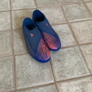 Blå och rosa Adidas Predator fotbollsskor - Säljer ett par blå och rosa Adidas Predator fotbollsskor. Skorna har en modern design med rosa detaljer och är perfekta för fotbollsträning och matcher. De är i bra skick och redo för nya äventyr på fotbollsplanen.