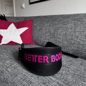 Svart träningsbälte från Better Bodies - Säljer ett svart träningsbälte från Better Bodies med rosa text. Perfekt för gymmet och ger bra stöd vid tunga lyft. Bältet har en justerbar spänne för optimal passform. Använt (se bild 4) men fungerar fortfarande perfekt. 