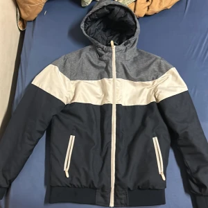 Blå och grå jacka från FSBN - Säljer en snygg blå och grå jacka från FSBN med huva och dragkedja. Jackan har vita detaljer och ribbade muddar. Perfekt för kyligare dagar.