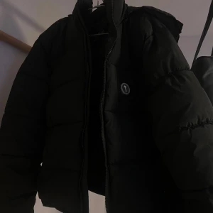 Svart pufferjacka från Trapstar - Säljer en svart pufferjacka från Trapstar med huva och dragkedja. Jackan har en broderad logga på ryggen och är perfekt för kyliga dagar. Den är både stilren och funktionell. Har en zipp på luvan också om man använda jackan utan luva. Har knappt används köpt nyligen men passar ej… trade off/byte funkar också.