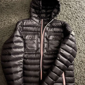 Svart pufferjacka från Moncler - .Snygg svart dunjacka från Moncler (köptes på plick) med huva och dragkedja i rött, vitt och blått. Jackan har två bröstfickor med knappar och ett quiltat mönster som ger en stilren look. Perfekt för kyliga dagar. Storlek 3 och fullt fungerande nfc scann. Hör av er vid fler frågor och funderingar🥂🙌