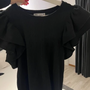 Svart topp med volang från Zara - Snygg svart topp från Zara med volangdetaljer på ärmarna. Perfekt för en stilren look. Tillverkad i ett mjukt material som ger en bekväm passform. ENDAST ANVÄND EN GÅNG!!