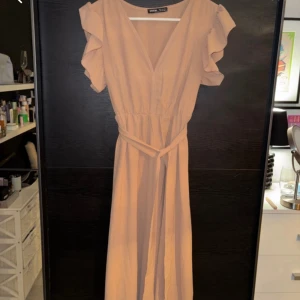 Beige långklänning med volangdetaljer - Elegant beige långklänning med v-ringning och volangdetaljer på ärmarna. Klänningen har ett knytband i midjan för en smickrande passform. Perfekt för sommarens festligheter.