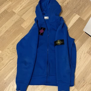 Blå hoodie från Stone Island - Säljer en snygg blå hoodie från Stone Island med dragkedja och huva. Den har märkets ikoniska patch på ärmen. Perfekt för en casual look.
