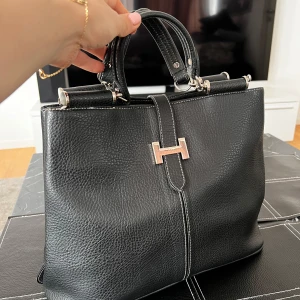 Svart handväska med metallspänne - Elegant svart handväska i skinn med ett stilrent metallspänne framtill. Väskan har två handtag och en strukturerad yta som ger en lyxig känsla. Perfekt för att bära dina dagliga nödvändigheter med stil.