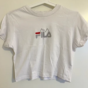 Vit croppad t-shirt från Fila - Säljer en vit croppad t-shirt från Fila med loggan i  grått och rött på framsidan. T-shirten har korta ärmar och en rund halsringning. Perfekt för en avslappnad stil. Bra skick! Kommer dock inte till användning längre❤️