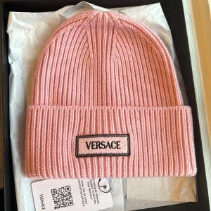 Rosa ribbad mössa från Versace - Snygg rosa mössa från Versace med ribbad design och broderad logotyp framtill. Perfekt för att hålla sig varm med stil. Den är helt oanvänd och slutsåld överallt. Originalpris ca 3000 kr🩷