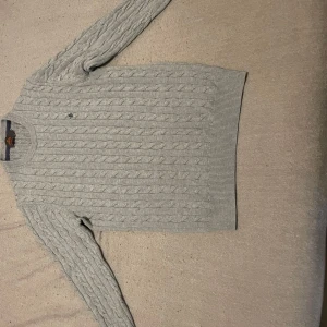 Grå kabelstickad tröja från Morris - Snygg grå kabelstickad tröja från Morris  med rund hals och långa ärmar. Perfekt för kyligare dagar med sin klassiska design och mjuka material.Tröjan är väldigt skön och är i nyskick