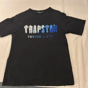 Svart t-shirt från Trapstar - Snygg svart t-shirt från Trapstar med tryck i vitt och blått på framsidan. Texten 'Trapstar It's a Secret' ger en cool look. Perfekt för en avslappnad stil.