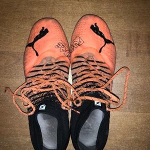 Orange och svarta fotbollsskor från Puma - Snygga fotbollsskor från Puma i orange och svart med snörning. Skorna har en modern design med Pumas logotyp och är perfekta för fotbollsplanen.
