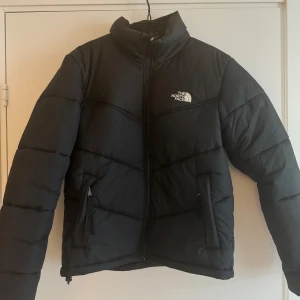 Svart dunjacka från The North Face - Säljer en snygg svart dunjacka från The North Face. Jackan har en dragkedja framtill och två praktiska fickor med dragkedjor. Perfekt för kyliga dagar med sin varma och vadderade design.