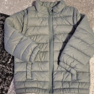 Grå quiltad jacka/väst från Kappahl (Ny) - Säljer en grå quiltad jacka från Kappahl i storlek 98. Jackan har en dragkedja framtill och avtagbara ärmar, vilket gör den mångsidig för olika väder. Perfekt för höst och vår!🌸😊 Den är helt ny och onavänd 