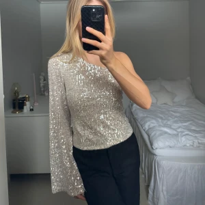 Glittrig oneshoulder topp - Snygg glittrig oneshoulder topp i silver med långa ärmar. Perfekt för nyår eller en festlig kväll. Toppen har en tight passform och är täckt av paljetter som ger en glamorös look.