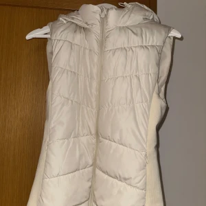 Beige dunväst från Zara - Säljer en snygg beige dunväst från Zara med dragkedja och huva. Perfekt för kyliga dagar när du vill hålla värmen utan att kompromissa med stilen. Västen har en quiltad design som ger en modern touch. säljes pga inte kommer till användning - slutsåld 