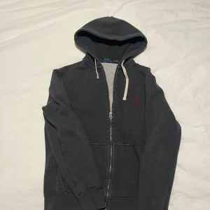 Grå hoodie från Polo Ralph Lauren - Säljer en stilren grå hoodie från Polo Ralph Lauren med dragkedja och huva. Den har en liten röd broderad logga på bröstet och vita snören vid huvan. Perfekt för en avslappnad stil. Storlek S