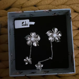 Silverfärgade blommiga örhängen från Ioaku - Eleganta silverfärgade örhängen från Ioaku med blomdesign. Örhängena har en kedjedetalj som ger en unik touch. Perfekta för att lyfta vilken outfit som helst. Örhängena är enbart använda en gång och är köpta för 600kr. Box ingår och örhängena decificeras innan de postas! Örhängena är ofta slutsålda och svåra att få tag på! 