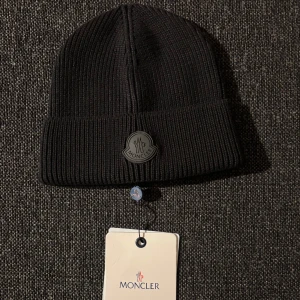 Svart ribbad mössa från Moncler - Snygg svart ribbad mössa från Moncler med broderad logotyp framtill. Perfekt för kyliga dagar och ger en stilren look. Passar både till vardag och mer uppklädda tillfällen.