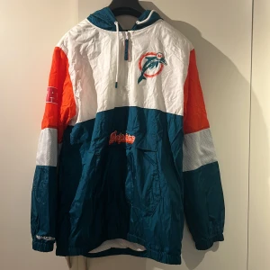 Flerfärgad vindjacka med logotyp - Säljer en cool flerfärgad vindjacka från Starter med Miami Dolphins logotyp. Jackan har en dragkedja framtill och en praktisk huva. Färgerna är en mix av blå, vit och orange, vilket ger en sportig look.