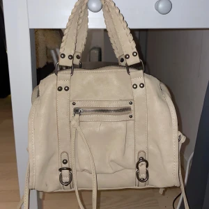 Beige handväska/axelväska  - Snygg beige axelväska i syntetmaterial med flätade handtag och dragkedjedetaljer. Väskan har flera fack och en praktisk storlek för vardagsbruk. Super fin och passar till nästan alla outfits!💕