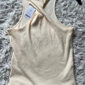 Beige najila tank från Gina Tricot - Säljer en snygg beige ribbad linnetopp från Gina Tricot. Toppen har en unik twistad design vid halsen och är ärmlös, perfekt för varma dagar. 
