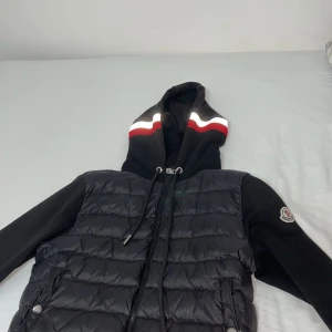 Svart jacka med huva från Moncler - Snygg svart jacka från Moncler med huva och dragkedja. Jackan har en quiltad design på framsidan och ärmar med Moncler-logga. Perfekt för kyligare dagar med stil.