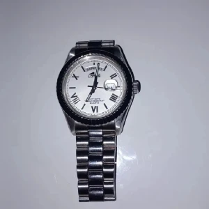 Silverfärgad klocka från Lotus - Snygg silverfärgad klocka från Lotus med romerska siffror och dag- och datumvisning. Den har en klassisk design med en robust metallarmband och en vit urtavla. Perfekt för en stilren look.