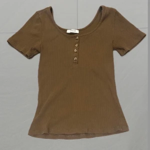 Khaki ribbad topp från Pieces - Säljer en khaki ribbad topp från Pieces med korta ärmar och knappar framtill. Perfekt för en avslappnad stil. Toppen är i storlek S och har en mjuk och bekväm passform.