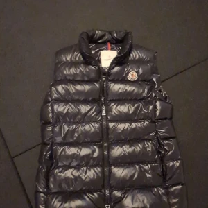 Svart dunväst från Moncler - Snygg svart dunväst från Moncler med dragkedja och hög krage. Perfekt för kyliga dagar när du vill hålla stilen. Västen har Monclers ikoniska logga på bröstet och en cool serietidningsdetalj på insidan.