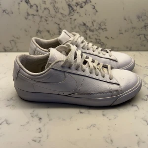 Vita sneakers från Nike - Säljer ett par klassiska vita Nike sneakers med snörning. Perfekta för en stilren look. Skorna har en enkel design med diskret Nike-logga på sidan och baksidan. Fräscha men har lätt användning på sig, har mindre orange märke på ett ställe vid Nike märket, se bild. 