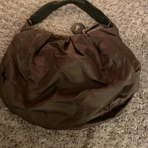 Brun axelväska från Lacoste - Snygg brun axelväska från Lacoste med ett klassiskt krokodilmärke. Väskan har en praktisk dragkedja och är perfekt för vardagsbruk. Den har en stilren design och är tillverkad i ett slitstarkt material.