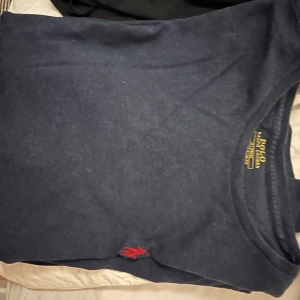 Mörkblå t-shirt från Polo Ralph Lauren - Snygg mörkblå t-shirt från Polo Ralph Lauren med en liten röd broderad logga. Den har en klassisk rund hals och är kortärmad. Perfekt för en stilren och avslappnad look.