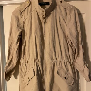 Beige jacka från Zara Basic - Snygg beige jacka från Zara Basic med dragkedja och knappar. Jackan har trekvartsärmar och praktiska fickor framtill. Perfekt för vår och höst!