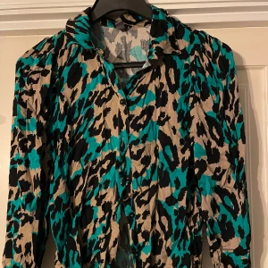 Mönstrad skjorta från Couture - Snygg skjorta med leopardmönster i turkos, beige och svart från Couture. Den har långa ärmar och knappar framtill. Perfekt för en djärv stil.