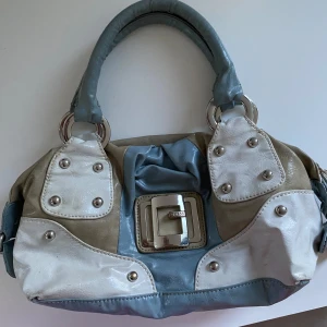 Vintage 2000s Guess Väska - Snygg handväska från Guess i flera färger som beige, blå och vit. Väskan har ett coolt patchwork-mönster med silverdetaljer och en praktisk dragkedja. Insidan är fodrad med ett färgglatt mönster och har en innerficka med dragkedja. 