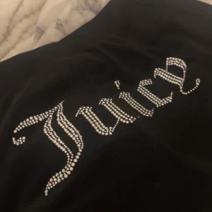 Svarta Juicy Couture byxor med strassdetaljer - Snygga svarta byxor från Juicy Couture med glittrande strassdetaljer. Byxorna har en bekväm passform och är perfekta för en avslappnad stil. Det ikoniska Juicy-logotypen pryder baksidan i strass. 2 kristaller är borta på bakdelen och en metall grej på snöret är borta men endå super bra skick