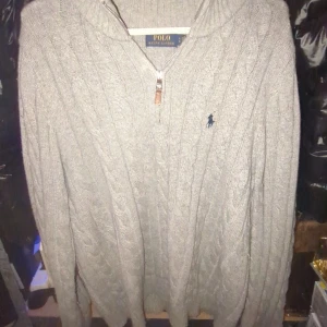 Ralph Lauren zip  - Säljer en stilren grå stickad tröja från Polo Ralph Lauren med dragkedja och broderad logga på bröstet. Tröjan har en klassisk kabelstickad design och långa ärmar. Perfekt för kyliga dagar! Typ helt ny använd själv 4 gånger ska inte va nå fläckar.XL i barn så mera som M L 