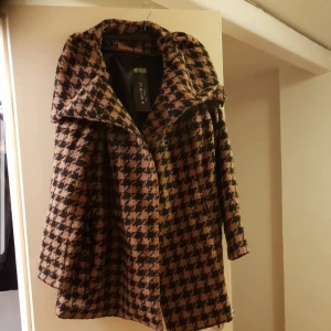 Rutig kappa från Aneau - Snygg rutig kappa från Aneau i svart och beige. Den har en stor krage och knappar framtill. Strl 40/42. Helt ny.