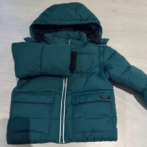 Grön pufferjacka med huva - Säljer en grön pufferjacka från Zara med huva och dragkedja. Jackan har långa ärmar och praktiska fickor framtill. Perfekt för kyliga dagar. Jackan för barn 24-36 månader helt ny!