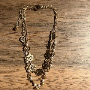 Guldarmband med pärlor och detaljer - Elegant guldhalsband med flera kedjor och dekorativa detaljer. Halsbandet har små vita pärlor och utsmyckade guldplattor som ger en lyxig känsla. Perfekt för att lyfta vilken outfit som helst.