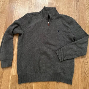 Grå stickad tröja från Ralph Lauren - Säljer en stilren grå stickad tröja från Ralph Lauren med dragkedja och ribbad krage. Perfekt för kyligare dagar med sin klassiska design och diskreta logga på bröstet.