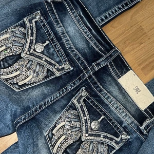 Blå Miss Me jeans med paljett och diamant detaljer  - Helt nya jeans från Miss Me med unika paljettdetaljer på bakfickorna. Jeansen har en bootcut-stil och är dekorerade med glittriga knappar och kontrastsömmar. Perfekta för att ge din outfit det lilla extra. Dom är i storlek 23 och i längd 32