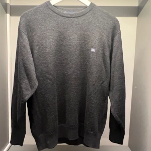 Grå tröja från Burberry - Säljer en stilren grå tröja från Burberry London. Tröjan har en diskret logga på bröstet och är perfekt för en klassisk look. Den är långärmad och har ribbade muddar vid ärmslut och nederkant.