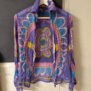 Färgglad sidenblus från Italien - Säljer en unik och färgglad blus i 100% siden, tillverkad i Italien. Blusen har ett psykedeliskt mönster i lila, rosa, blått och gult. Den är långärmad med knappar framtill och en klassisk krage. Perfekt för dig som vill sticka ut!