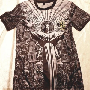 Fotboll t-shirt Limited edition. med Kristusmotiv - Unik t-shirt med ett iögonfallande grafiskt tryck av Kristusstatyn omgiven av detaljerade mönster. Finns i två färgvarianter: en i svart och grått och en i svart och gult. Båda har korta ärmar och ett emblem med texten 'Brasil' på bröstet.