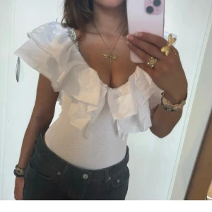 Vit ribbad topp med volangdetaljer - Säljer en stilren vit ribbad body med stora volangdetaljer vid axlarna. Toppen har en djup v-ringning och är ärmlös, vilket ger en elegant look. Perfekt för att lyfta vilken outfit som helst. Storlek S skriv om ni vill ha mer bilder