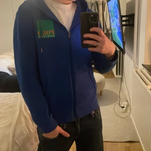 Blå hoodie från Polo Ralph Lauren - Säljer en snygg blå hoodie från Polo Ralph Lauren med dragkedja och huva. Den har en grön och gul logotyp på bröstet och ryggen. Väldigt unik tröja!