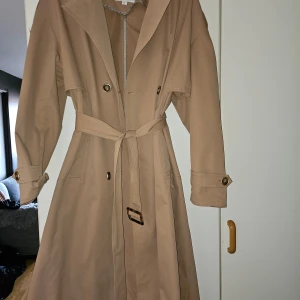 Beige trenchcoat från ZIAI - Snygg beige trenchcoat från ZIAI i storlek XL. Jackan har ett klassiskt bälte i midjan och knappar framtill. Perfekt för höst och vår med sin stilrena design och långa ärmar.
