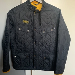 Svart quiltad jacka från Barbour - Säljer en snygg svart quiltad jacka från Barbour International. Jackan har en klassisk design med tryckknappar och dragkedja framtill. Perfekt för höst och vår!